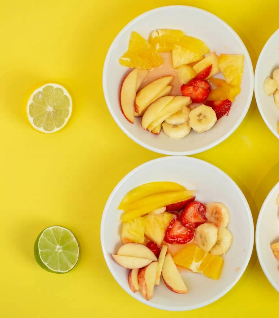 Two bowls of fruit salad. One with half a lemon on the side and one with a lime on the side